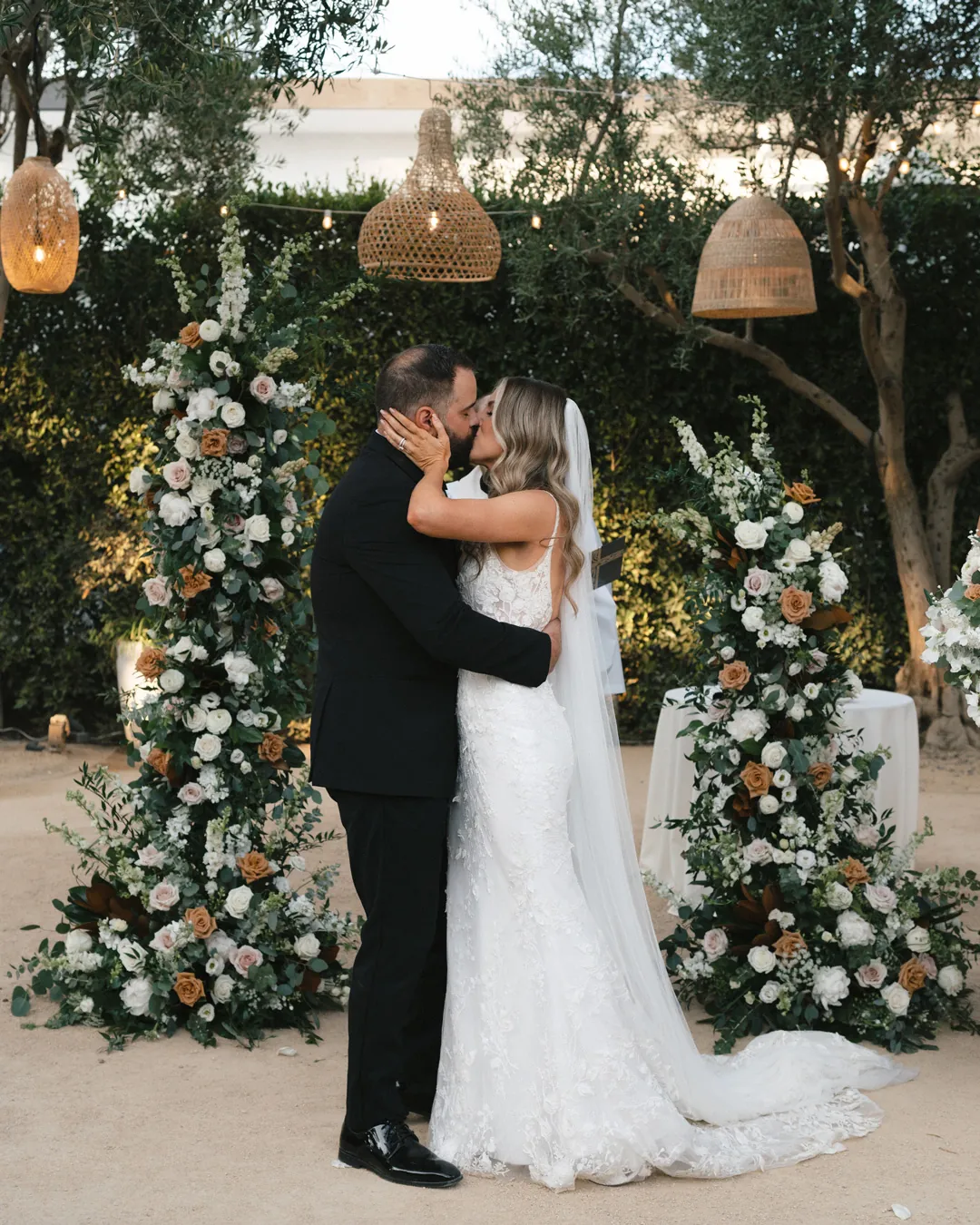 The couple kisses between two tall ceremony flower arrangements.