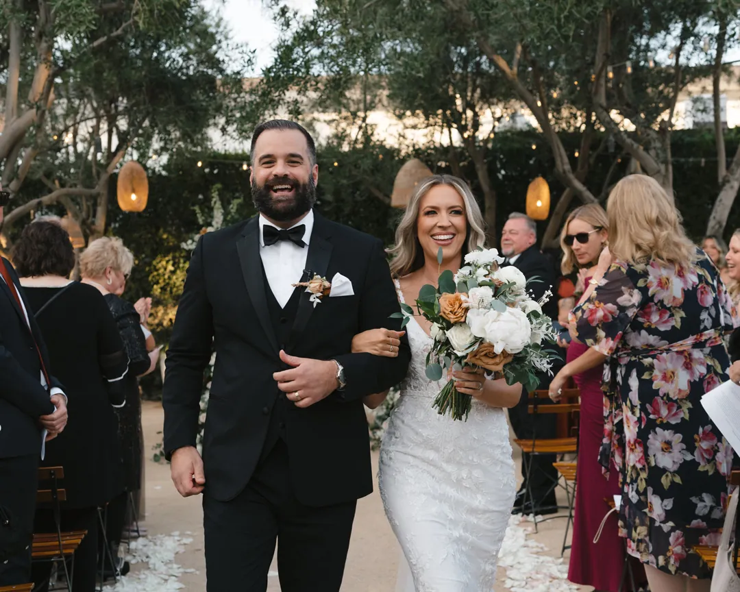 The couple walks together among smiling ceremony guests.
