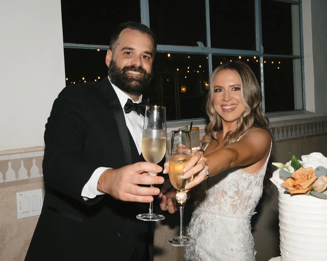 Newlyweds raising champagne glasses beside their wedding cake.