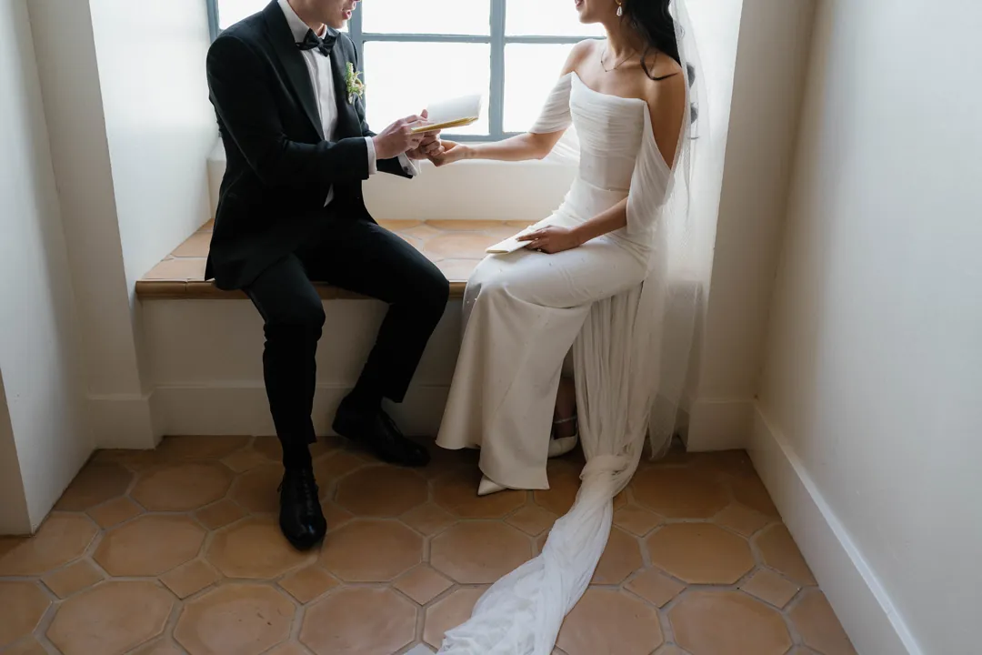 Bride and groom holding hands beside a sunlit window