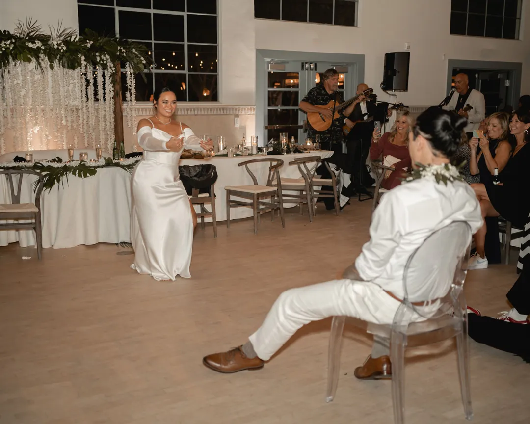 The bride stands on the dance floor while the groom sits nearby.