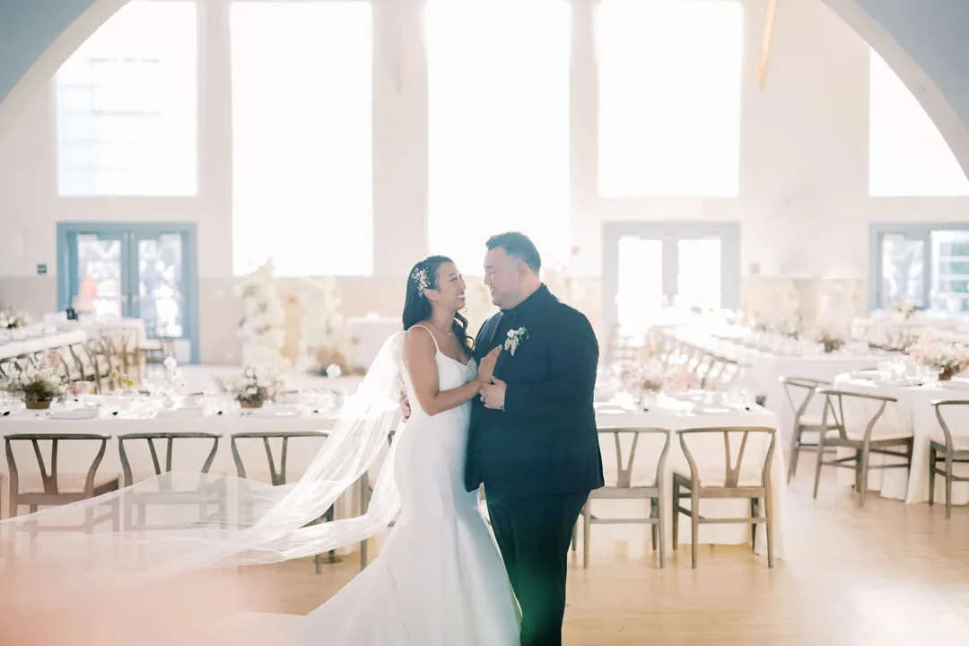 The couple embraces beside white linen reception tables.