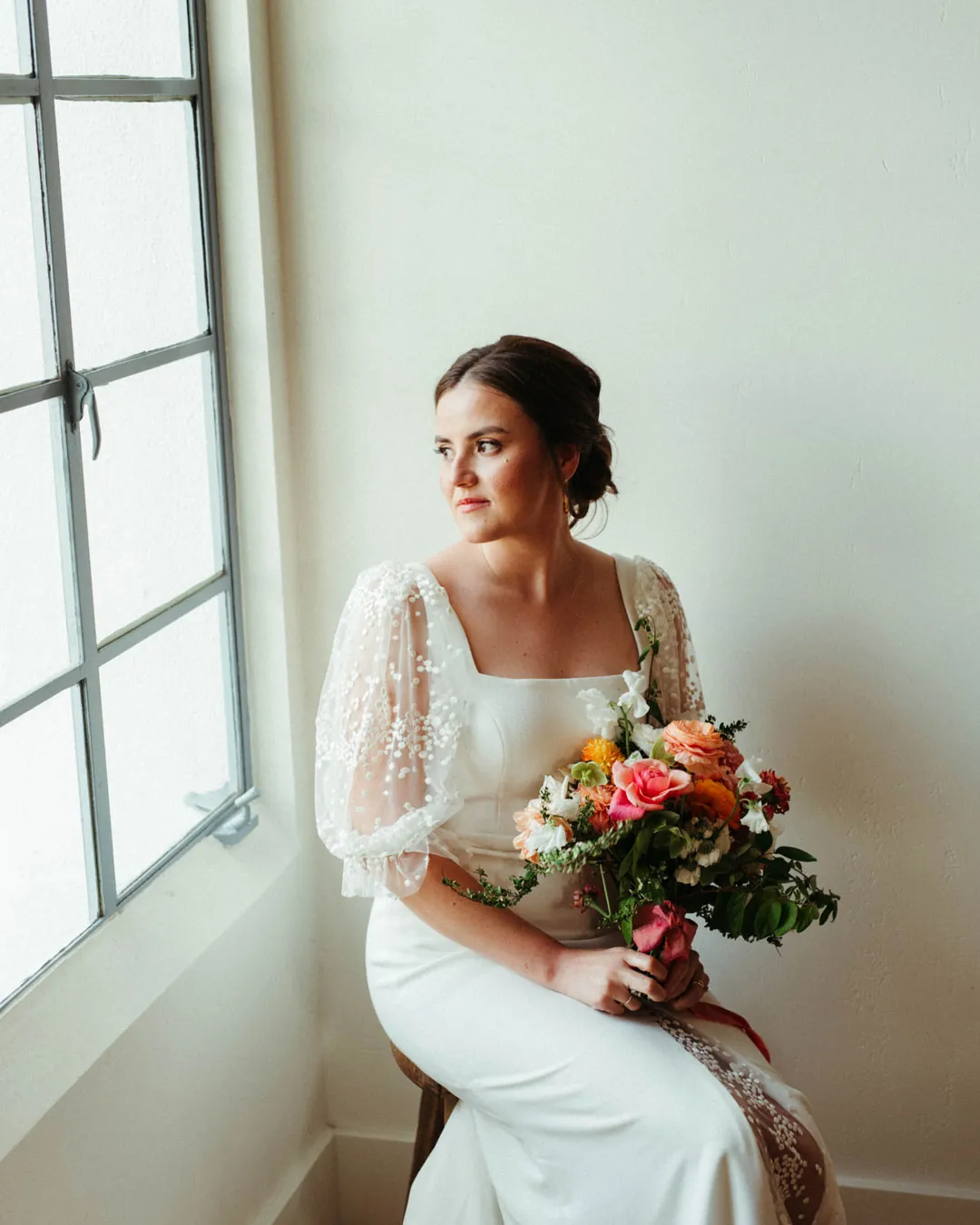 The bride holds a bouquet beside a bright window.