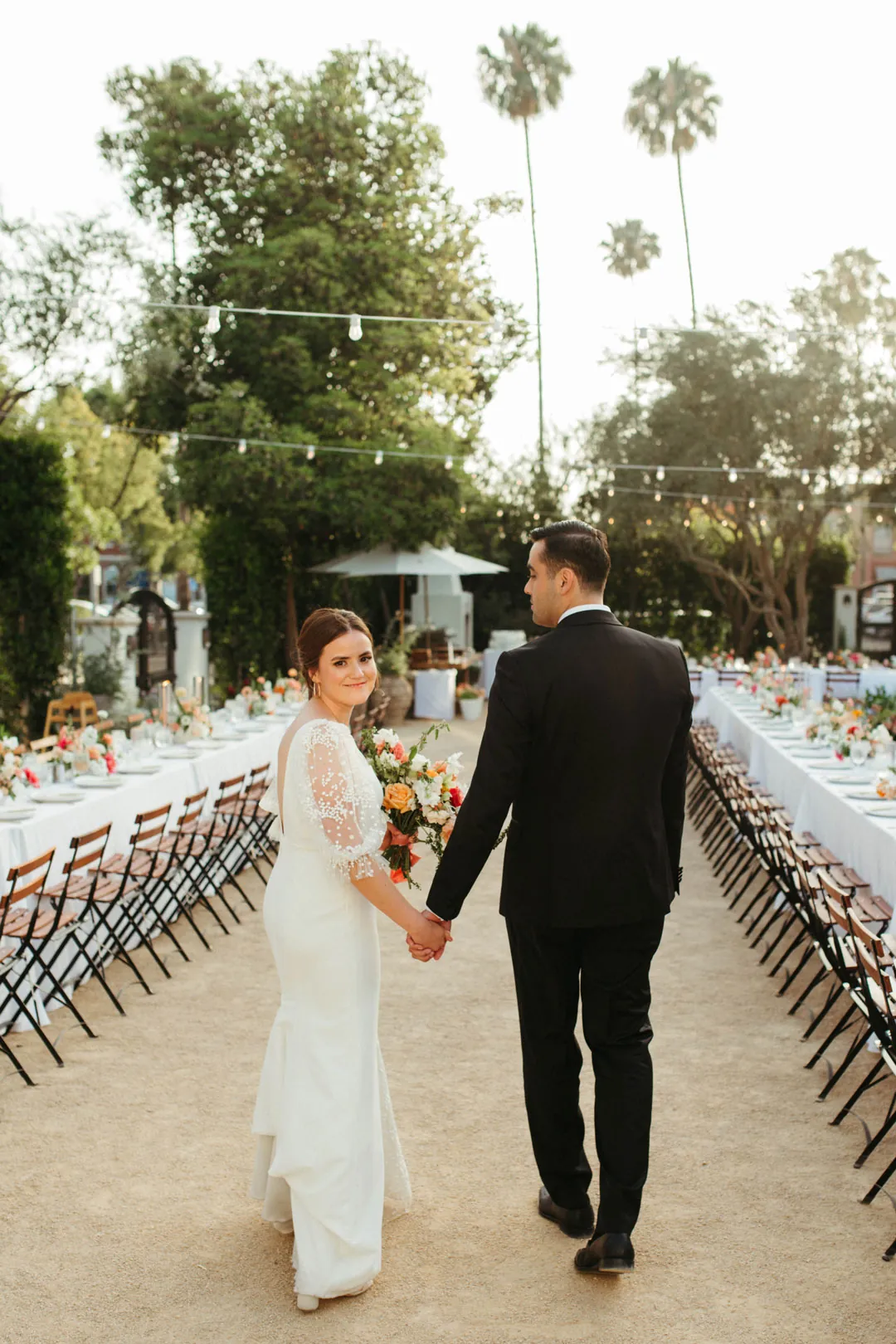The couple holds hands beside long courtyard reception tables.