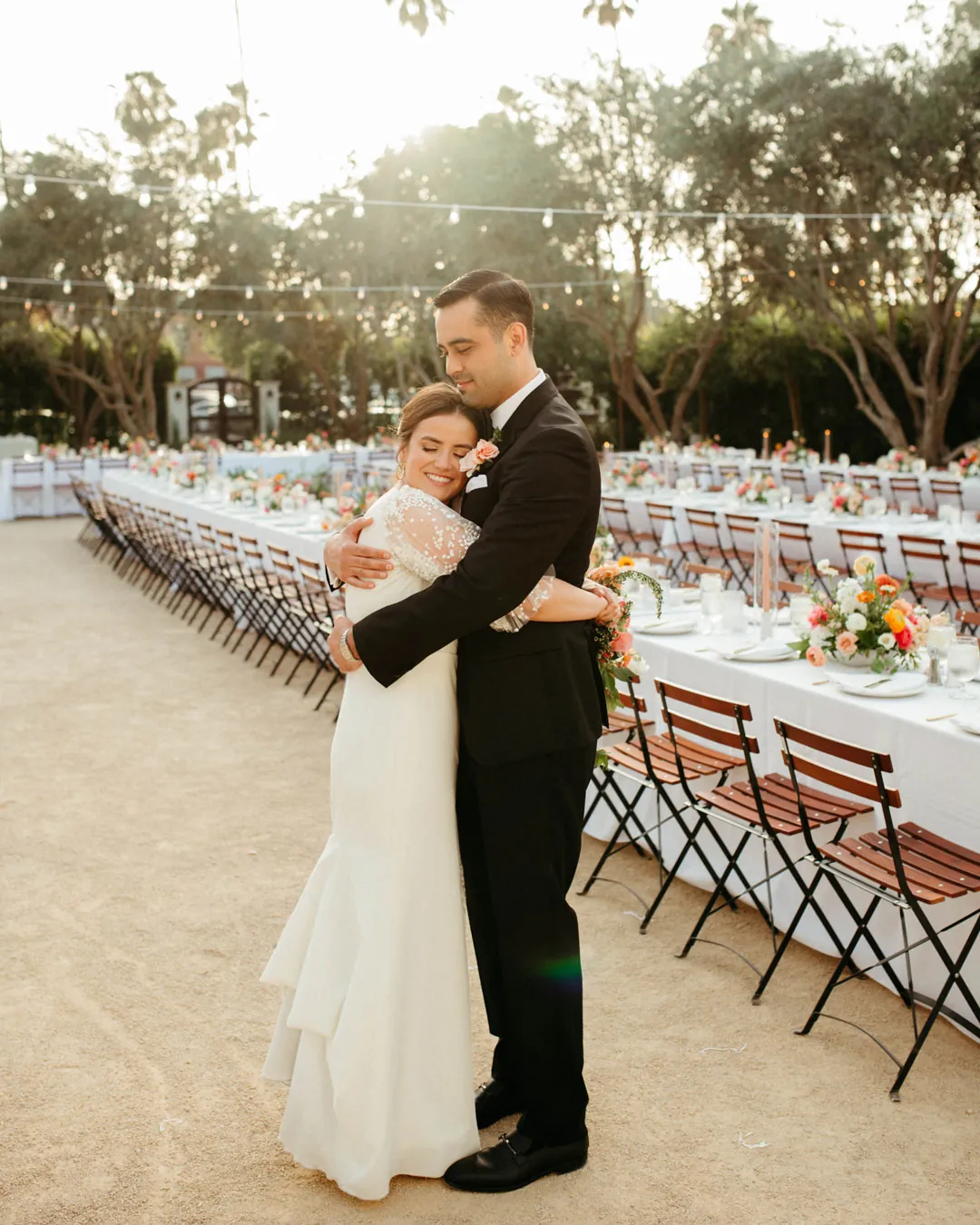 The couple embraces beside long outdoor reception tables.