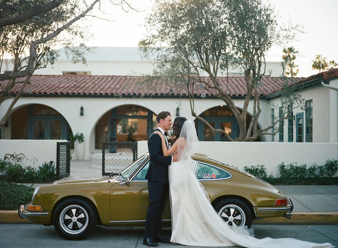Newlyweds kissing beside a vintage car in the courtyard.