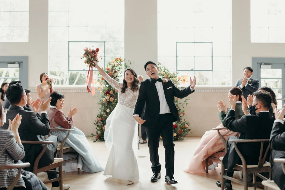 Newlyweds walking down the indoor aisle with raised arms.