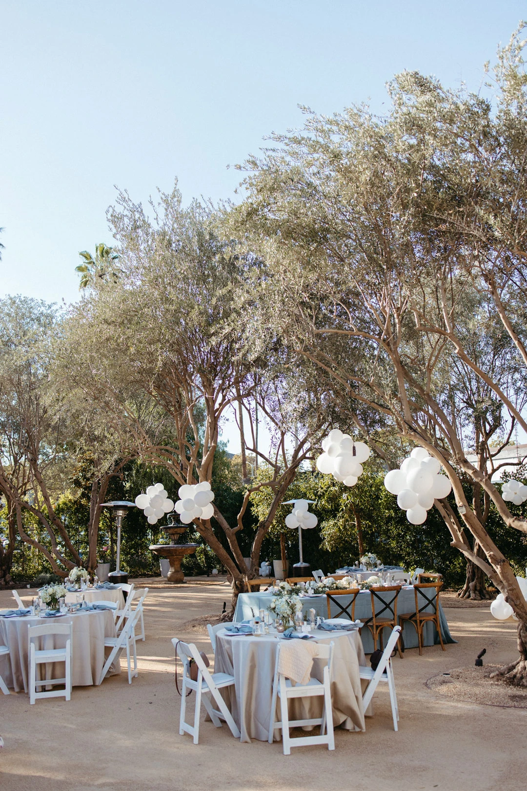 Round outdoor tables beneath trees and white balloons.