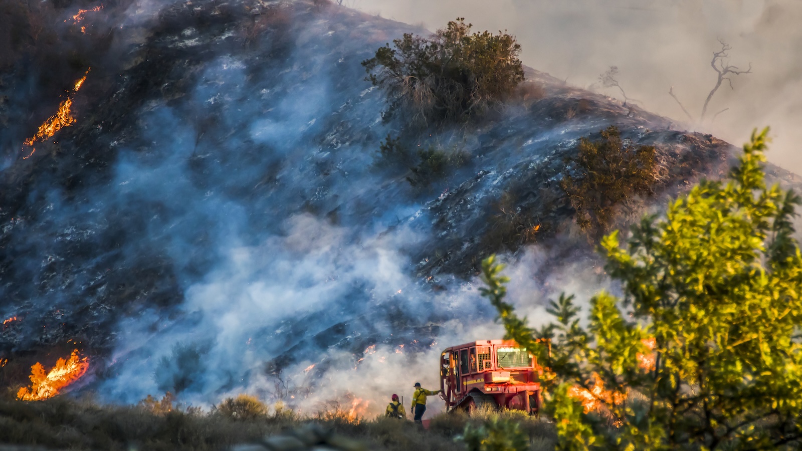 Two firefighters stand alongside a bulldozer as the California Woolsey Fire burns on the hill behind them.