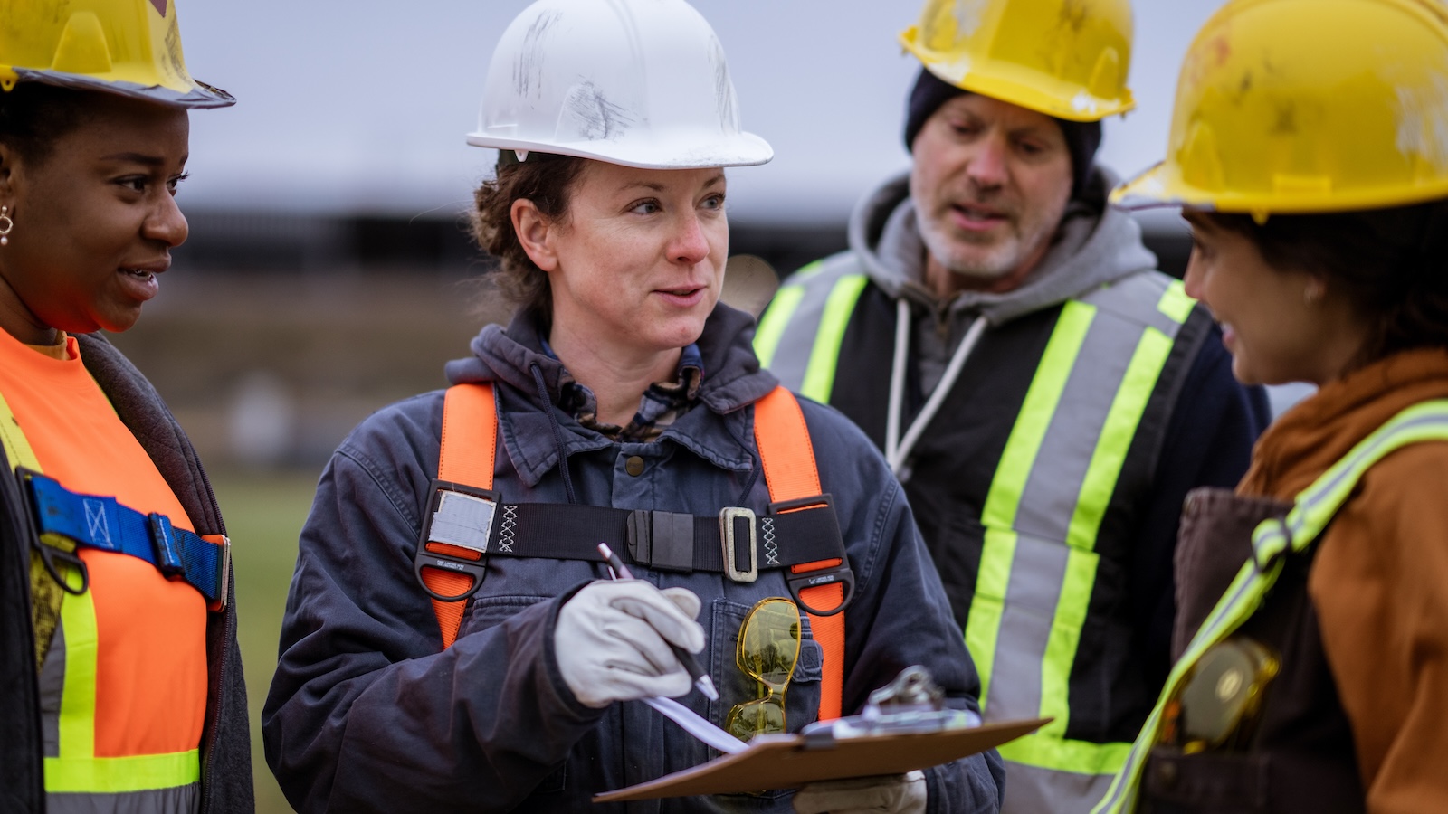 A crew chief in a hard hat and safety vest carries a clip board and speaks to members of her crew.