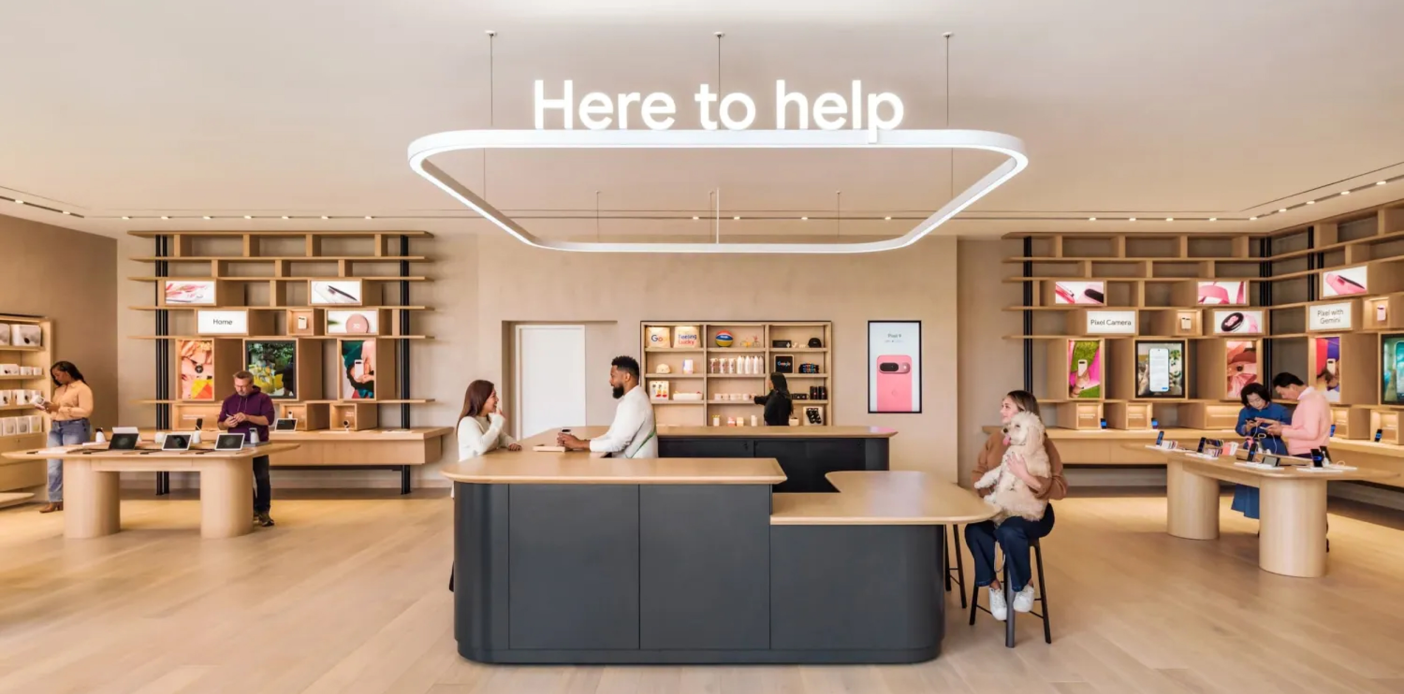 Modern retail store interior with customers interacting at wooden counters under a large lighted sign reading 'Here to help'.