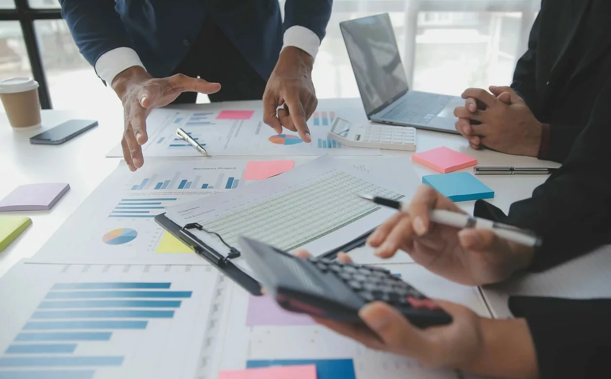 Trois personnes consultant des documents, une calcultracie utilisée et un ordinateur portable.