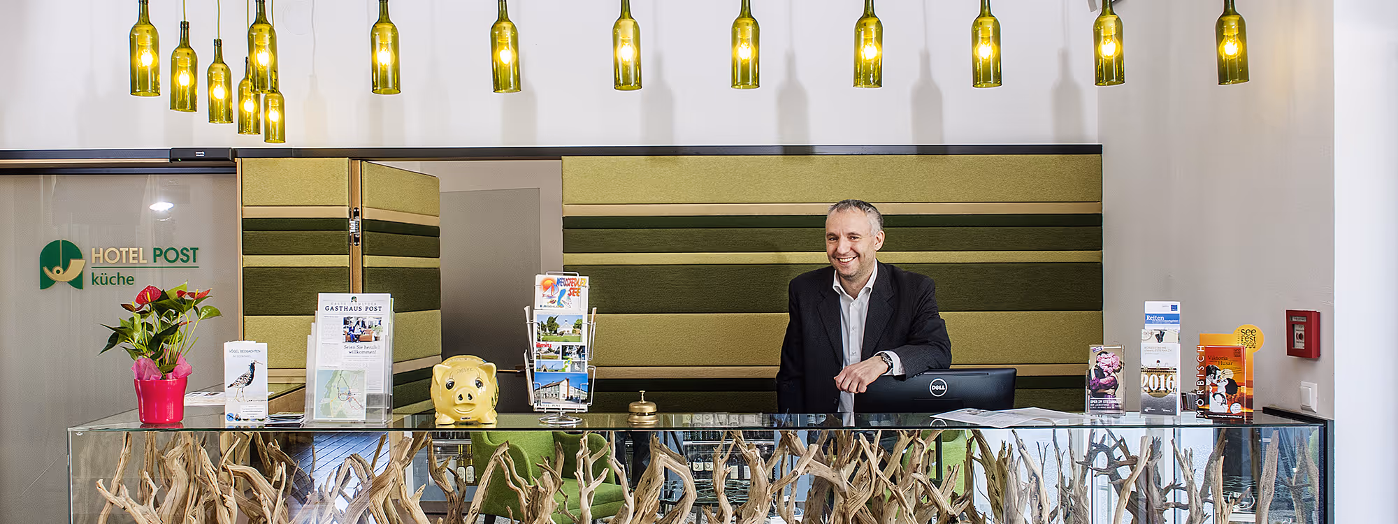 Hotelbetreiber Otto Egermann, der hinter einem mit Treibholz verzierten Glastresen steht, mit Broschüren und einem rosa Blumentopf auf dem Schreibtisch, Hotelrezeption im Hintergrund.