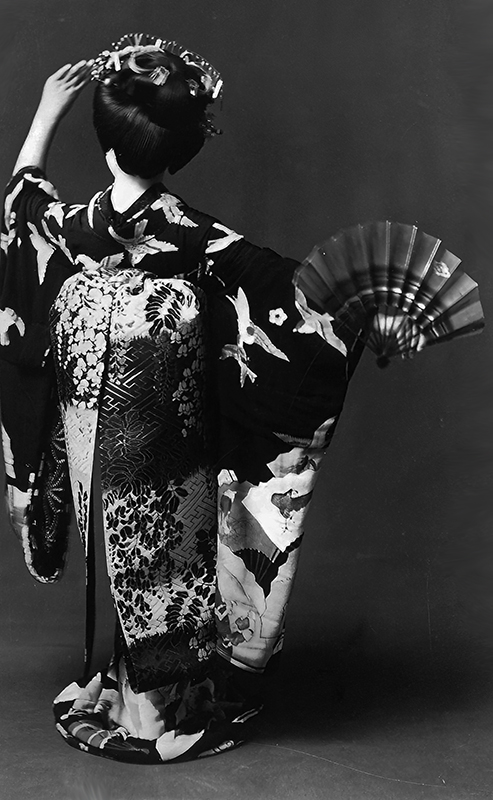 Geisha in traditional kimono holding folding fan, viewed from behind