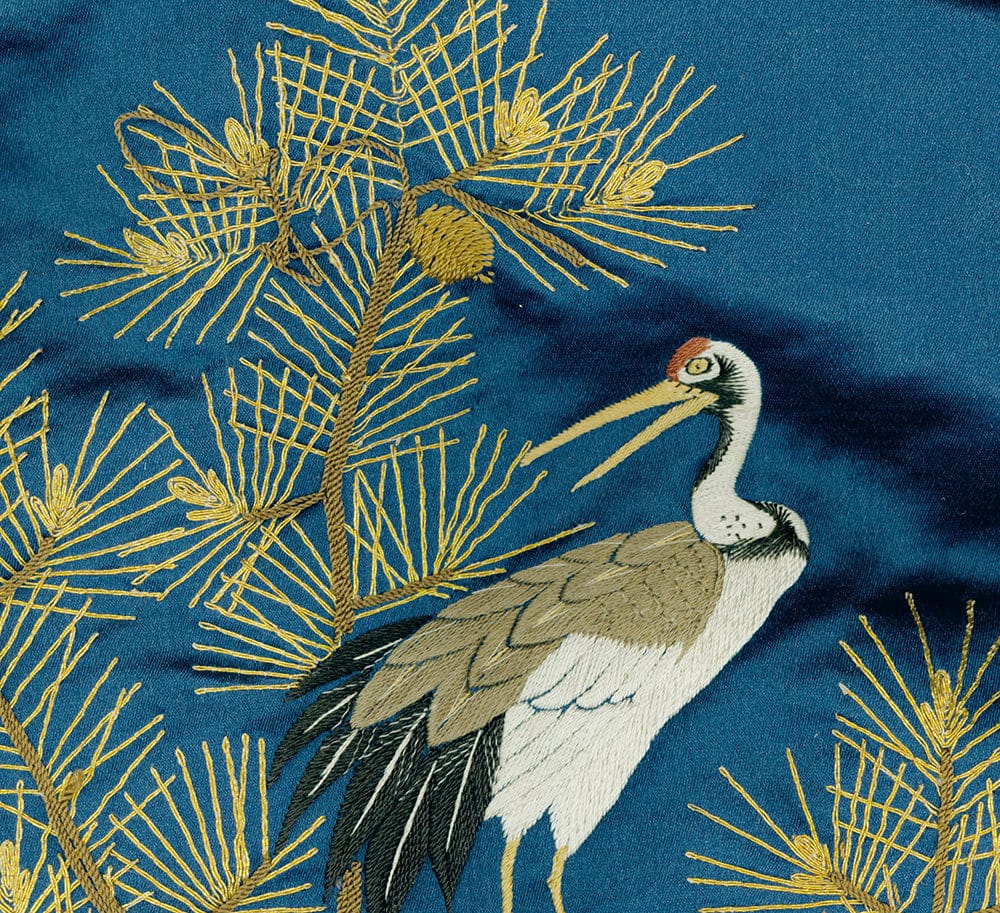 Embroidered crane standing among golden pine branches on blue fabric