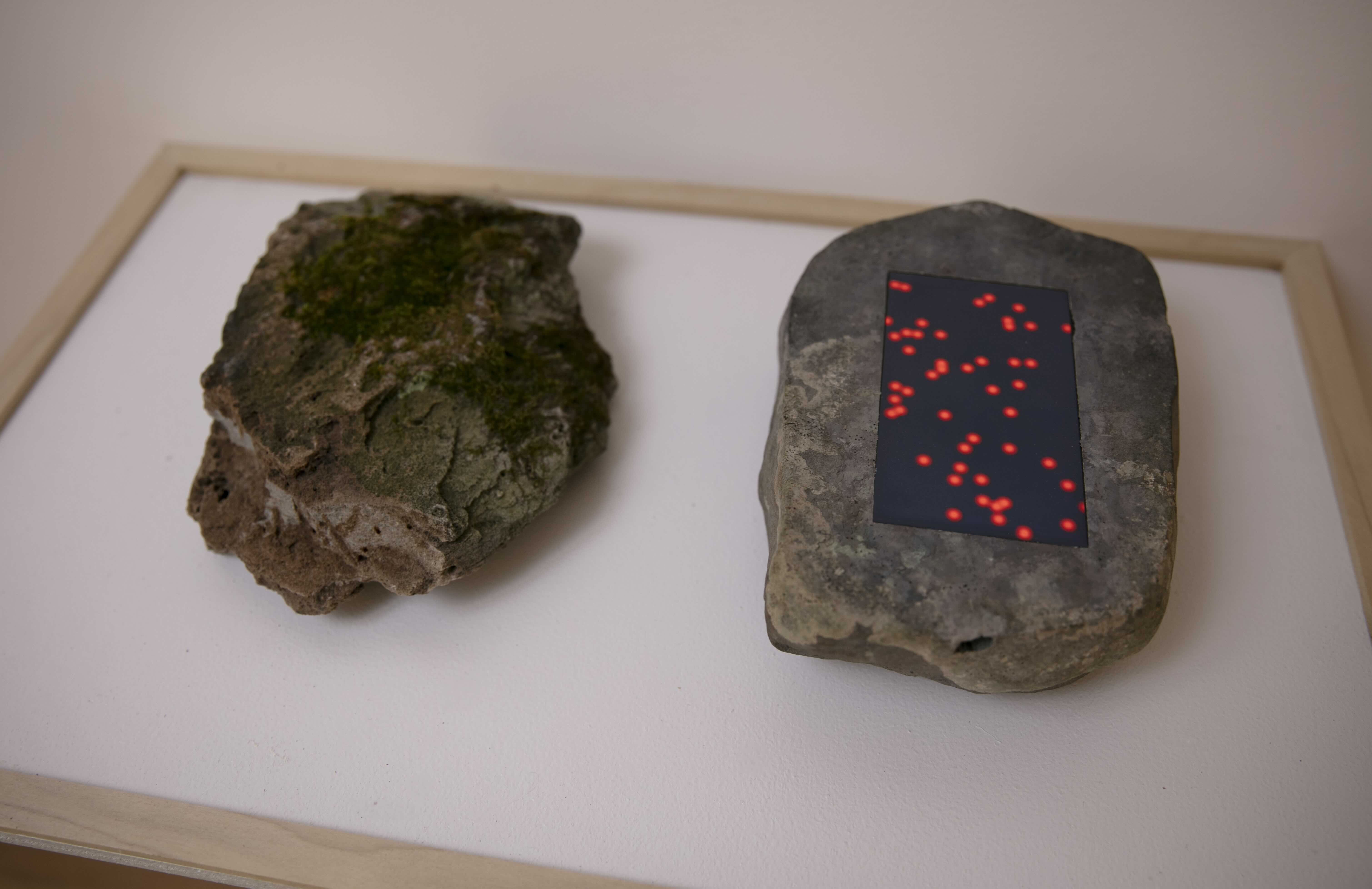 Artwork. Stone on a shelf with an inlaid screen showing red dots. Another stone next to it face down.