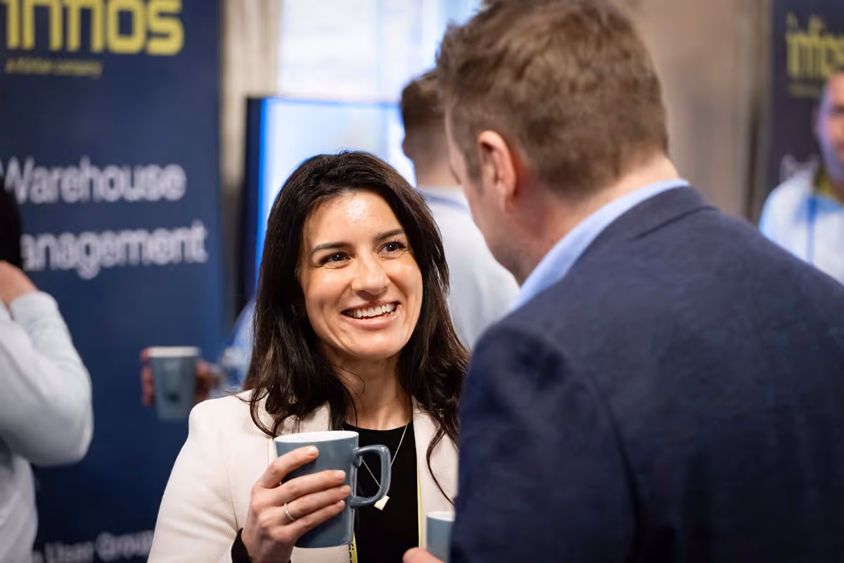 Professional corporate event photography from the company product launch, capturing a smiling female delegate holding a coffee cup while networking with a colleague in front of warehouse management brand banners.