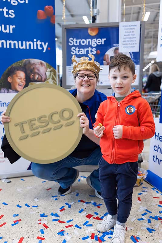 Professional PR event photography from the Tesco Stronger Starts Golden Grants initiative at the Dudley Extra store, capturing a staff member in a gold crown holding a large token prop next to a young participant amidst scattered floor confetti.