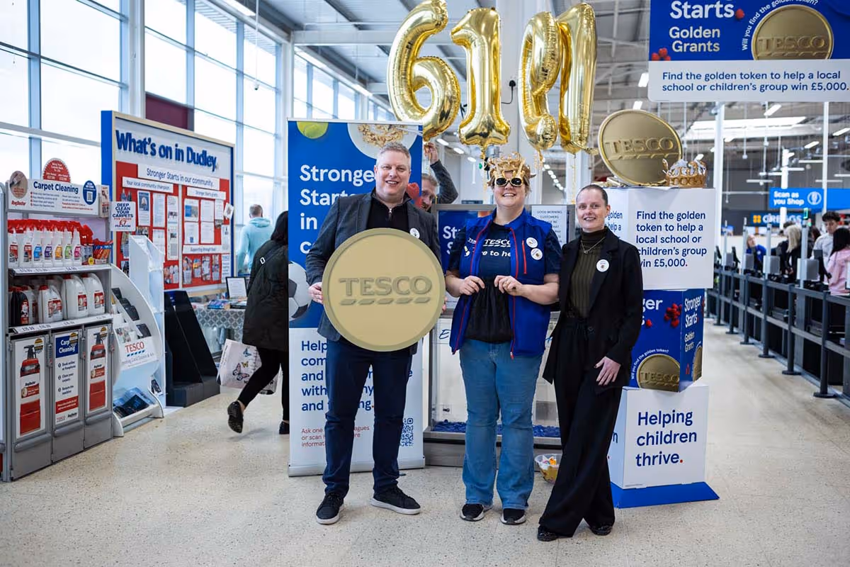 Commercial public relations photography from the Tesco Stronger Starts scheme at the Dudley Extra store, showing a corporate team standing with a large gold token display, promotional signage, and giant gold numeric balloons inside the supermarket foyer.