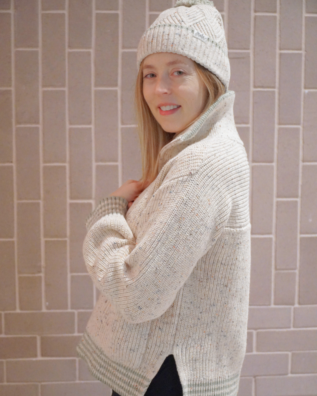 Pregnant woman wearing a cream-colored knit sweater with green trim and a matching knit hat with a pom-pom, standing sideways against a light brick wall, smiling gently.