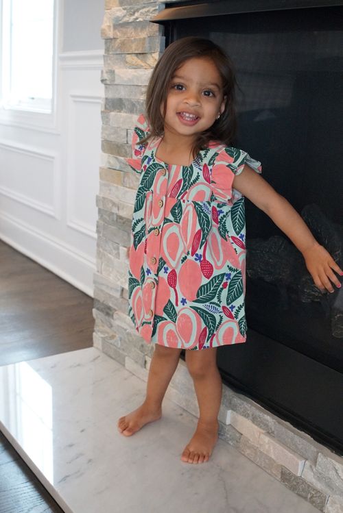 A young girl with dark hair stands barefoot on a marble hearth in front of a stone fireplace, smiling and wearing a colorful dress with a fruit and leaf pattern in pink, green, and red.