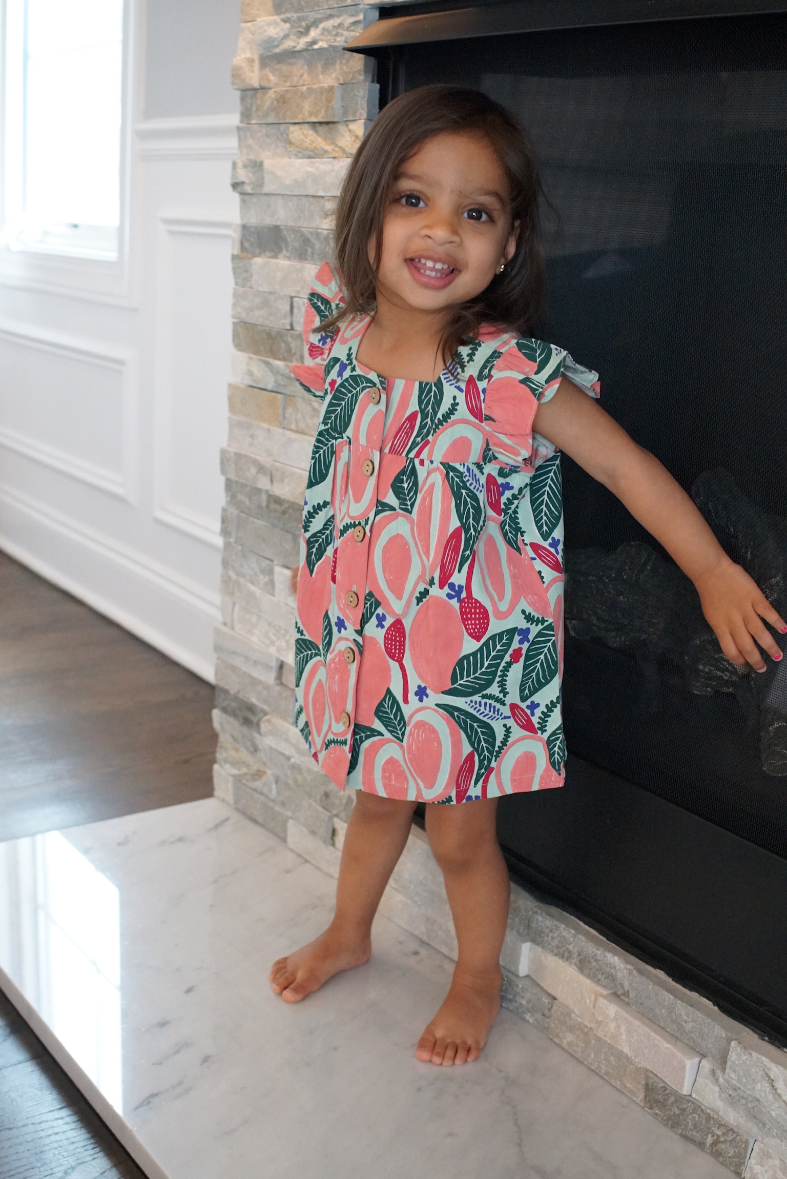 A young girl with dark hair stands barefoot on a marble hearth in front of a stone fireplace, smiling and wearing a colorful dress with a fruit and leaf pattern in pink, green, and red.