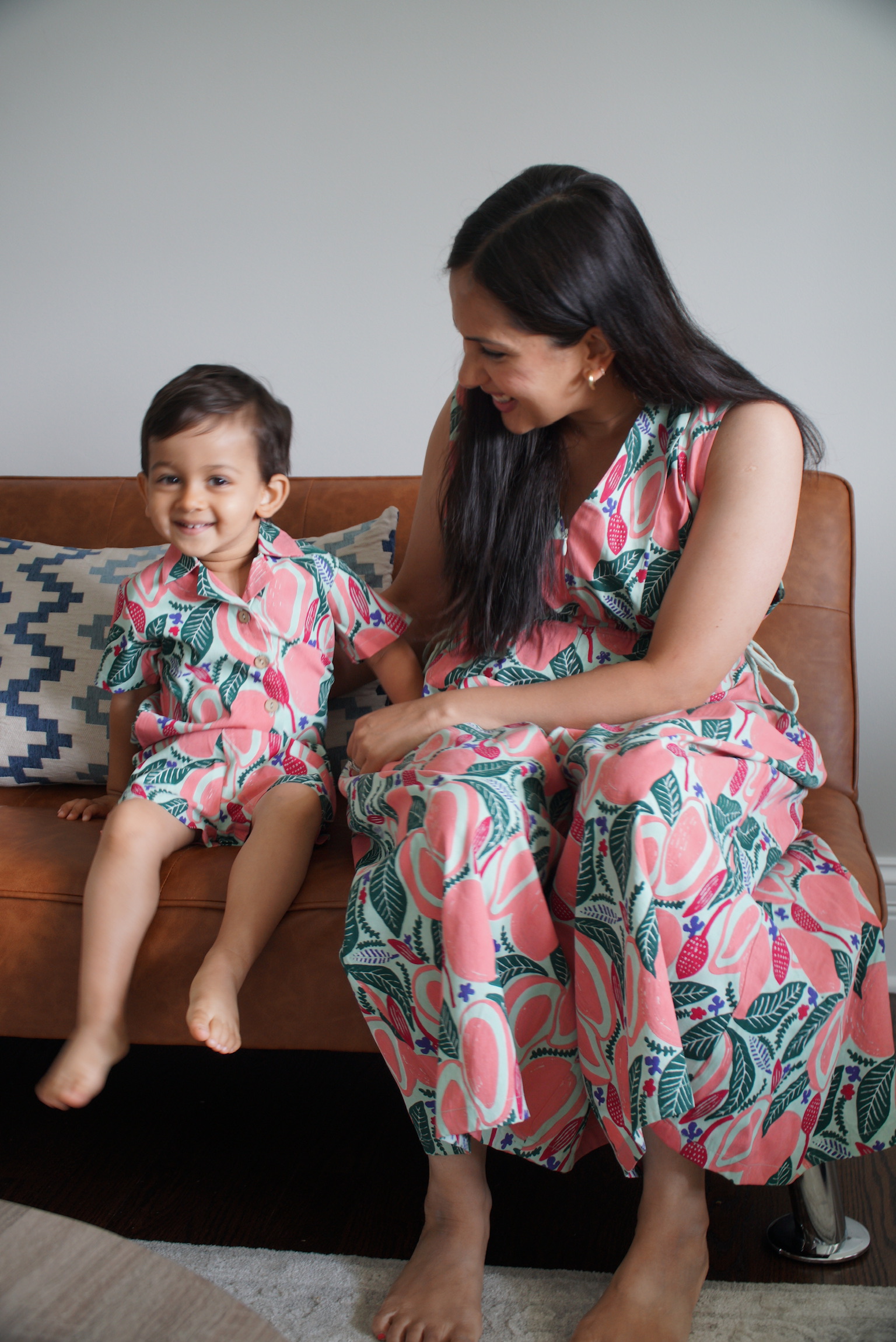 A woman and a young boy sitting on a brown leather couch, both wearing matching pink floral patterned outfits, the woman smiling at the boy who is looking towards the camera.