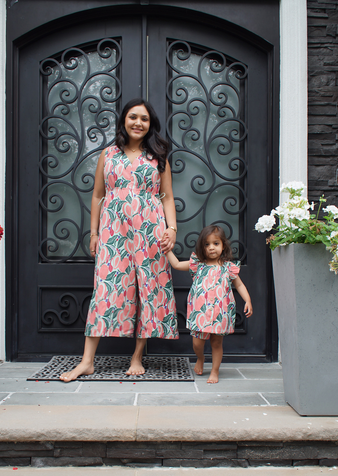 Woman and young girl standing barefoot on a stone porch in front of a black wrought iron decorative door, holding hands and wearing matching pink and green floral-patterned outfits.