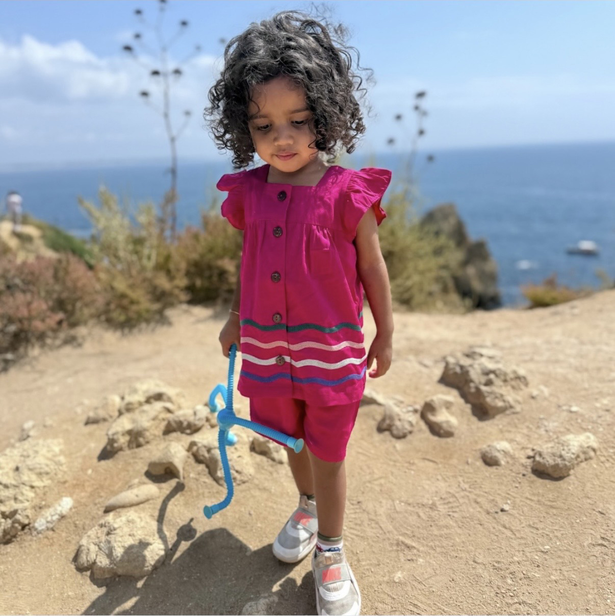 Young girl with curly hair wearing a magenta dress with striped trim and white sneakers stands on rocky dirt near the ocean, holding a blue rope toy and looking down.