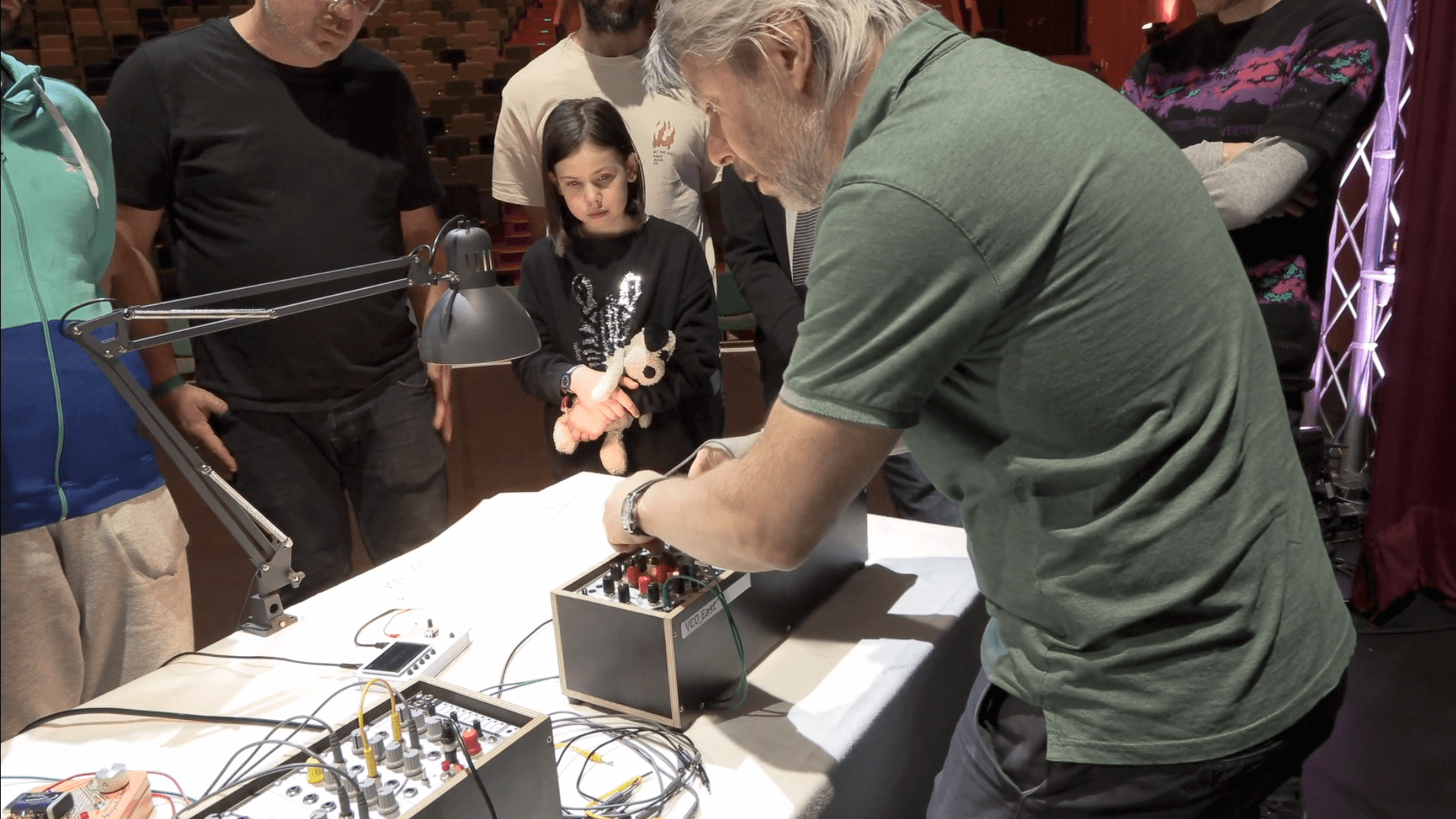 Ein Mann spielt auf einem Synthesizer, während ein Mädchen und andere Zuschauer ihm bei der Arbeit zusehen.