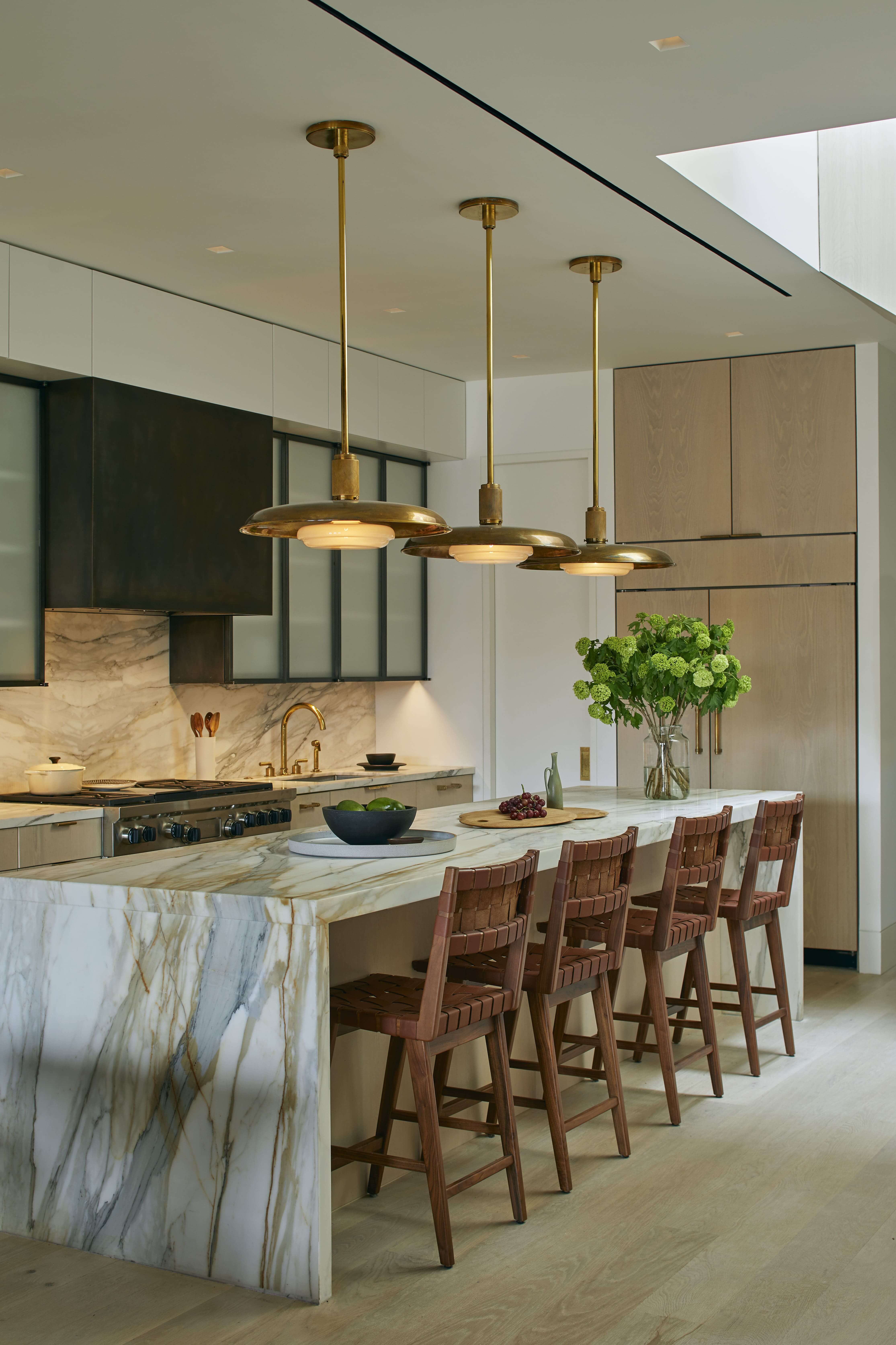 Modern kitchen with marble island, four wooden bar stools, brass pendant lights, and green flower centerpiece.
