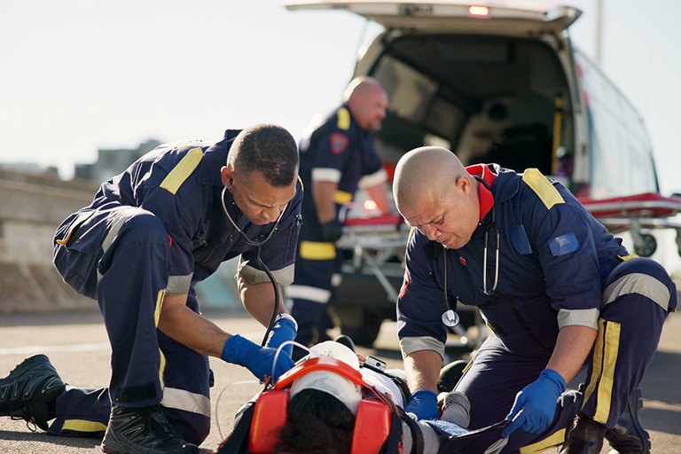Two first responders attend to someone on a bridge, while a third gets equipment out of a vehicle behind them.
