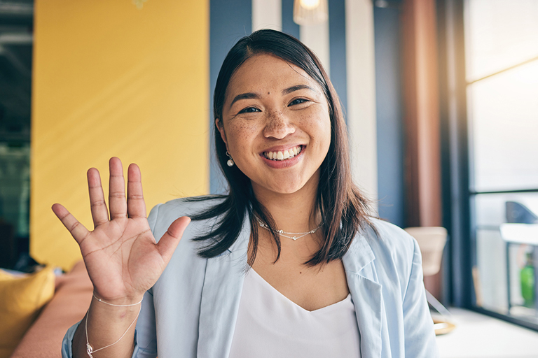 A young woman in a sunny office smiles and waves at the camera.