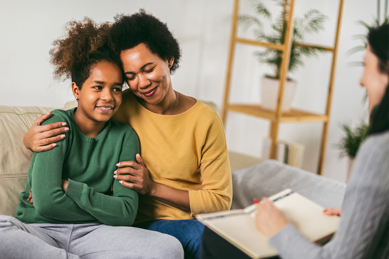 A mother and daughter happily embrace on a couch while a counsellor sits across from them with a notepad.