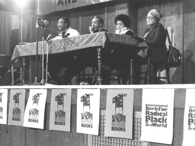 A black and white photo of the first International Book Fair of Radical Black and Third World Books. The photo is of a table on a stage with four people sitting and addressing an audience. From left to right: Norman Beaton, John La Rose, Yvonne Brewster and Pearl Connor