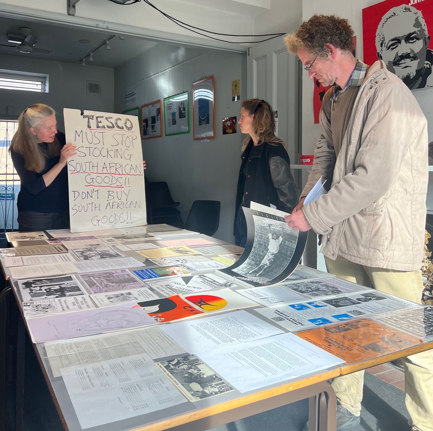 GPI Archivist is standing in the main room  holding a placard that reads, 'Tesco must stop stocking South African goods!! Don't buy South African Goods'. Two of the attendees are in the photo and are looking at the placard and other materials on display on a table