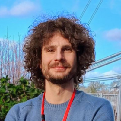 Close-up outdoor headshot of a person with curly hair wearing a blue top and red lanyard, standing in front of greenery and overhead cables against a clear sky.