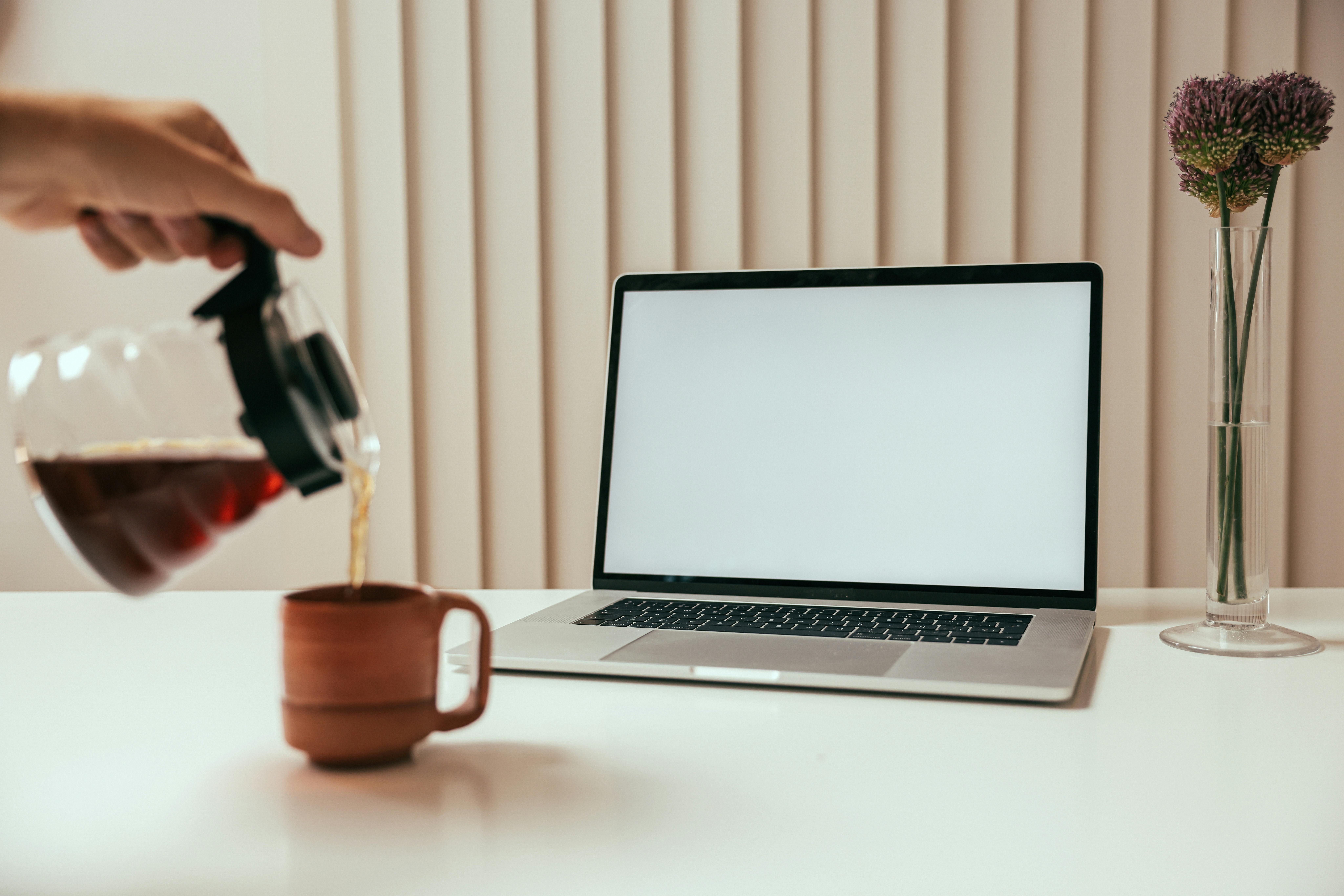 A clean workspace setup with coffee being poured beside a laptop and flower vase.