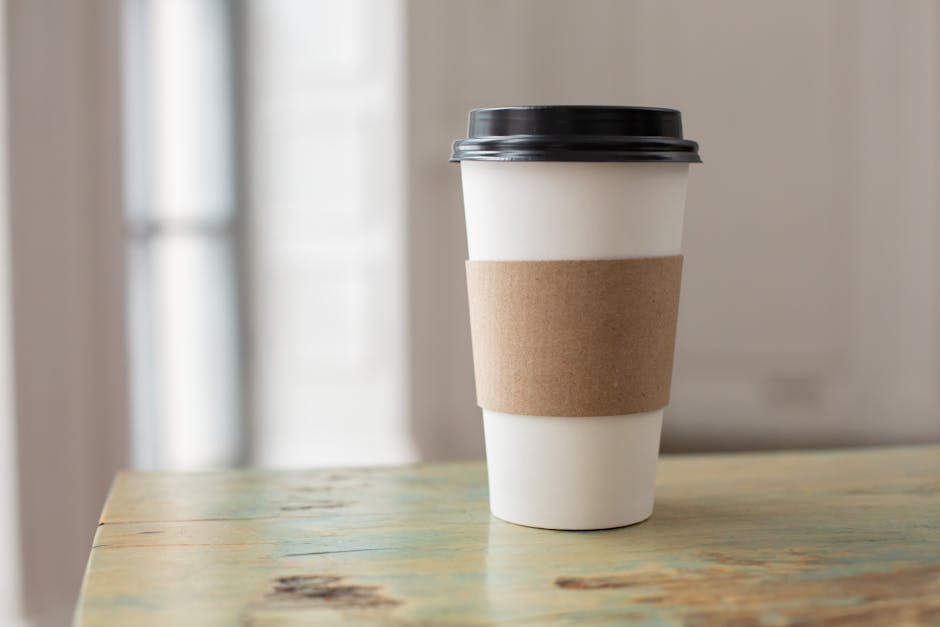 White paper coffee cup with brown cardboard sleeve on a rustic table indoors.