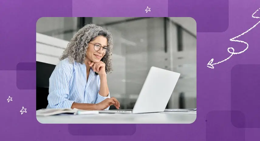 Smiling woman with curly gray hair working on a laptop at a desk.