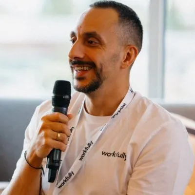 Man with short hair and beard speaking into a microphone, wearing a white Workfully t-shirt and lanyard.