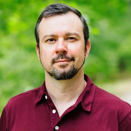 Sean Dazet with short brown hair and a beard, wearing a red shirt, smiling against a natural green background of trees