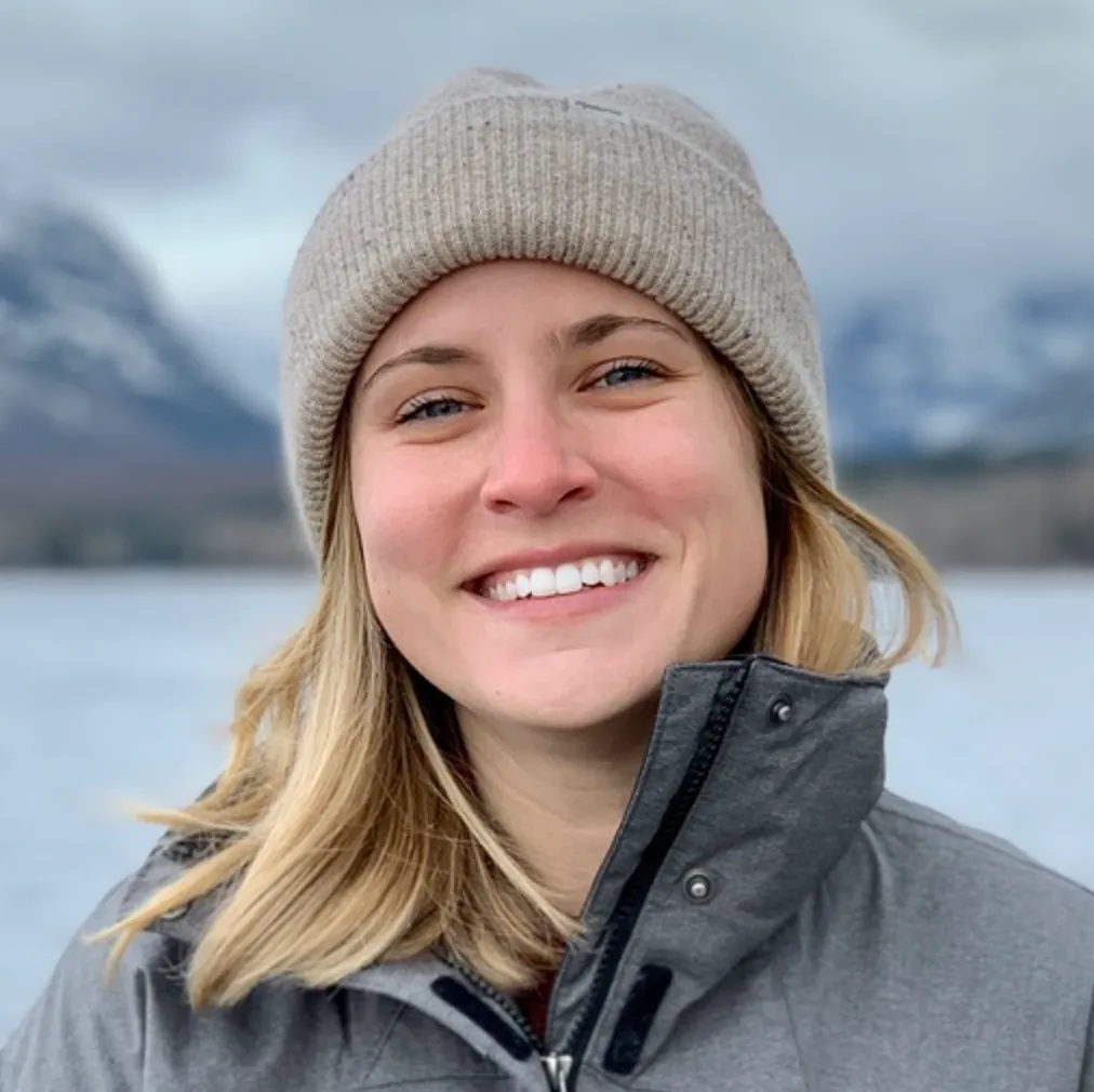 Smiling woman wearing a beige knit beanie and gray jacket with a snowy mountain and lake background.