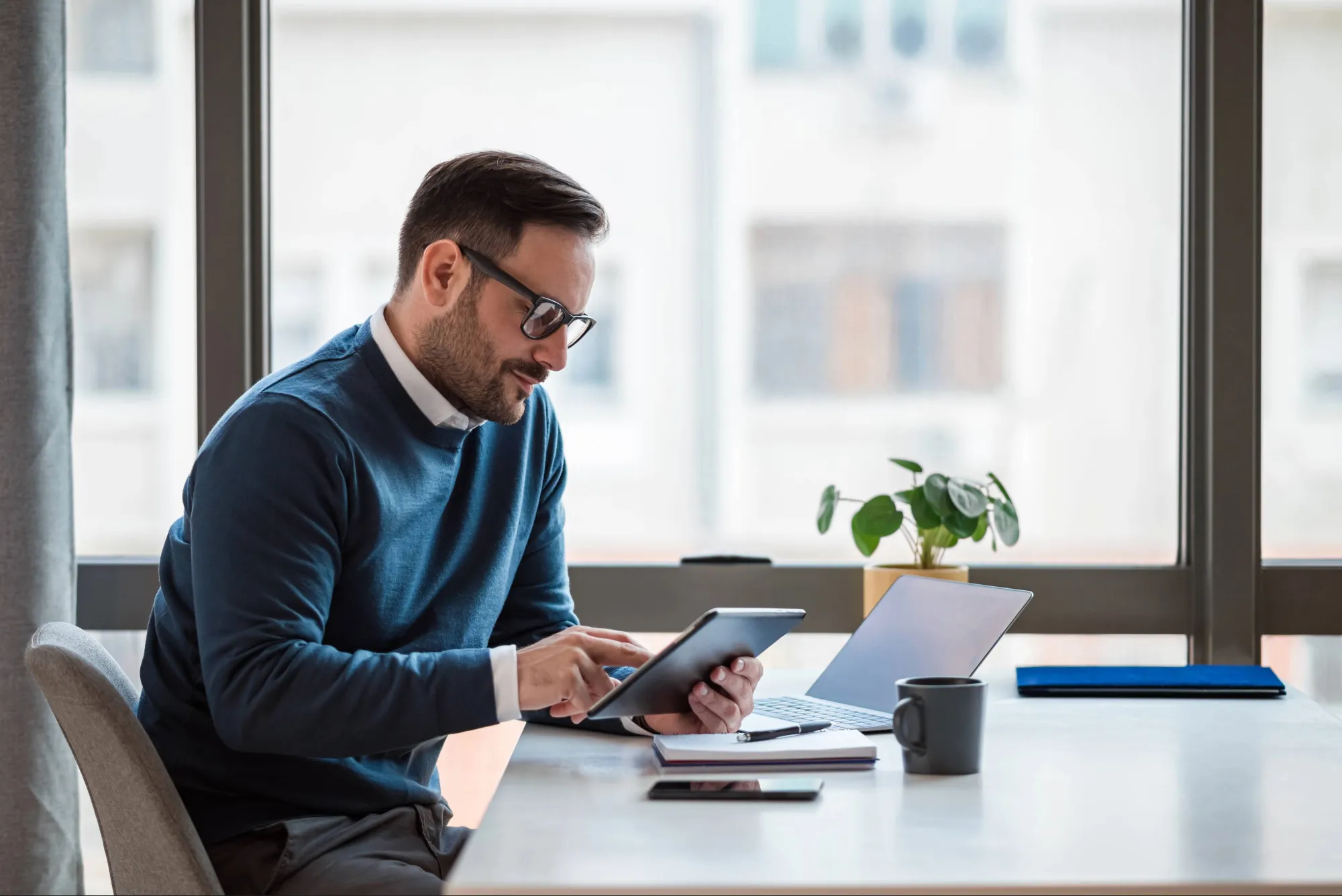 Businessman using digital tablet while working on laptop. Male executive with wireless computer at desk. He is sitting by window in office.