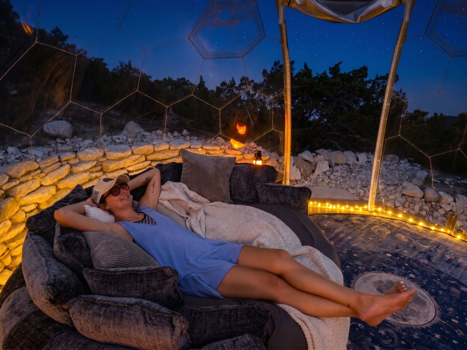 A guest lying back on the round daybed inside the Hype dome at dusk, limestone wall and cedars behind