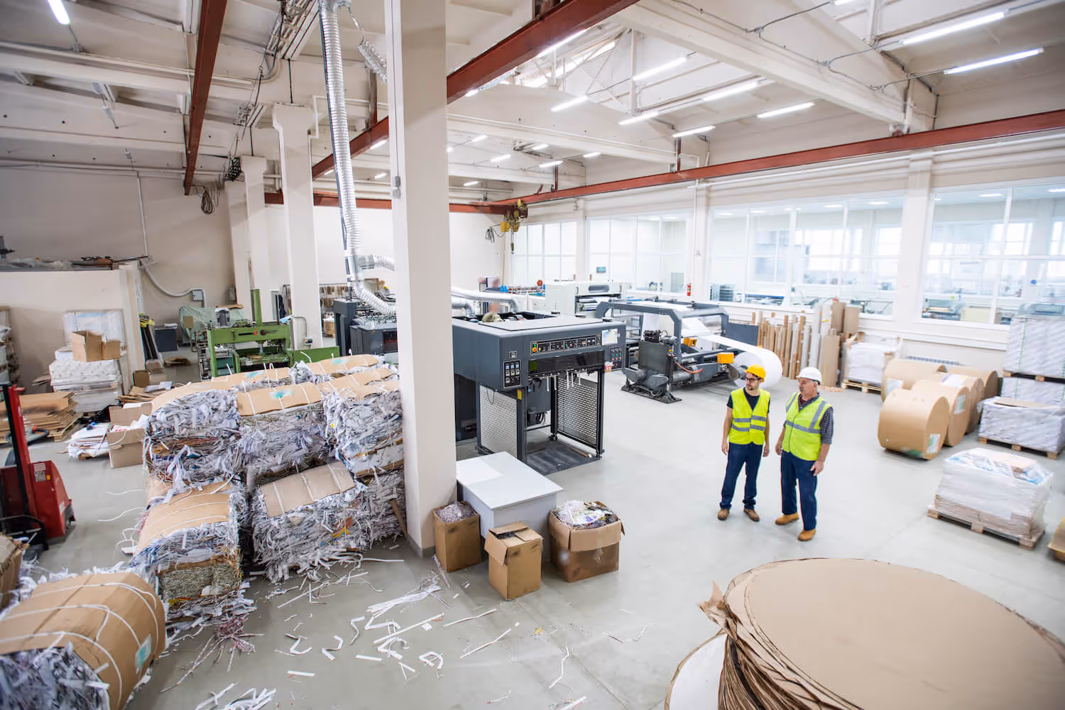 Two workers wearing safety vests and helmets standing in a spacious paper recycling factory surrounded by bales of shredded paper and industrial machinery.