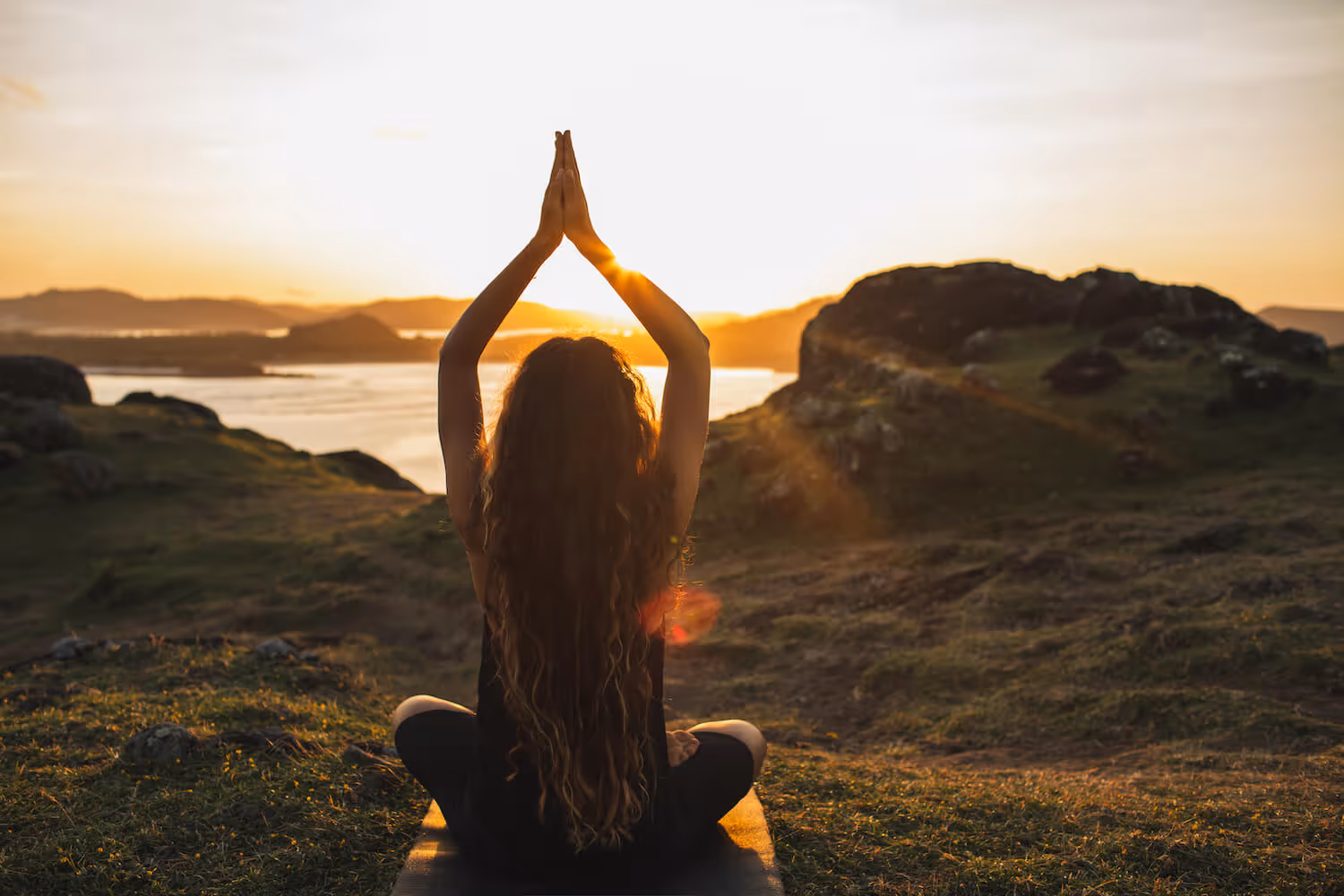 Woman with long hair practicing yoga meditation on a mat outdoors at sunset by a lake and hills.