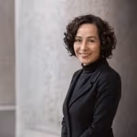 Smiling woman with short curly hair wearing a black turtleneck and blazer against a gray wall background.