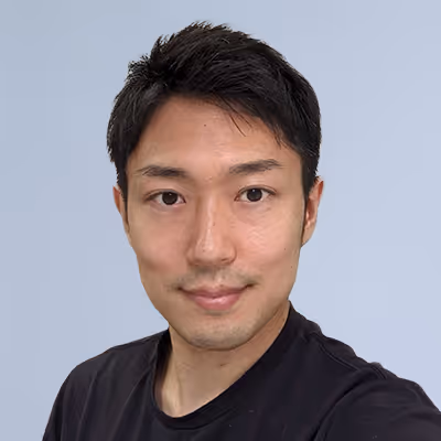 Close-up of a young man with short black hair wearing a black shirt against a plain light blue background.