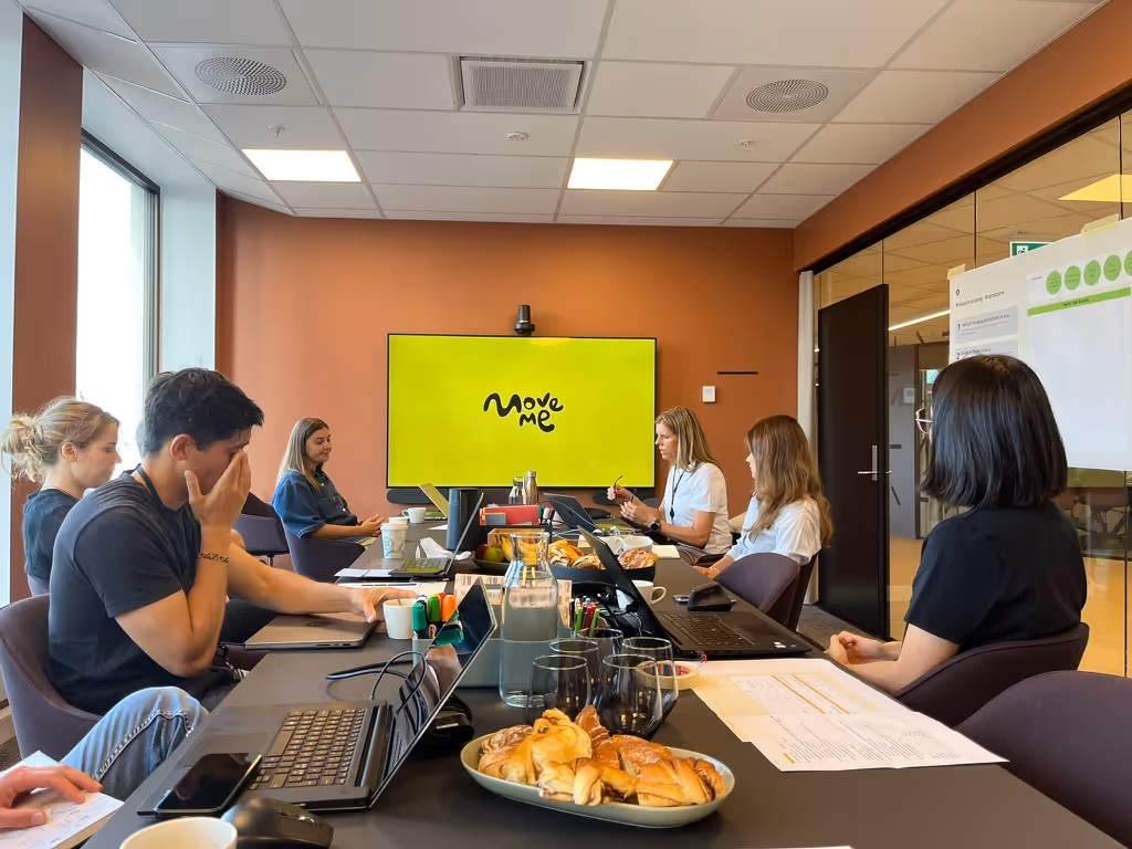 7 personer som har workshop i et møterom, med en stor MoveMe logo på skjermen i bakgrunnen